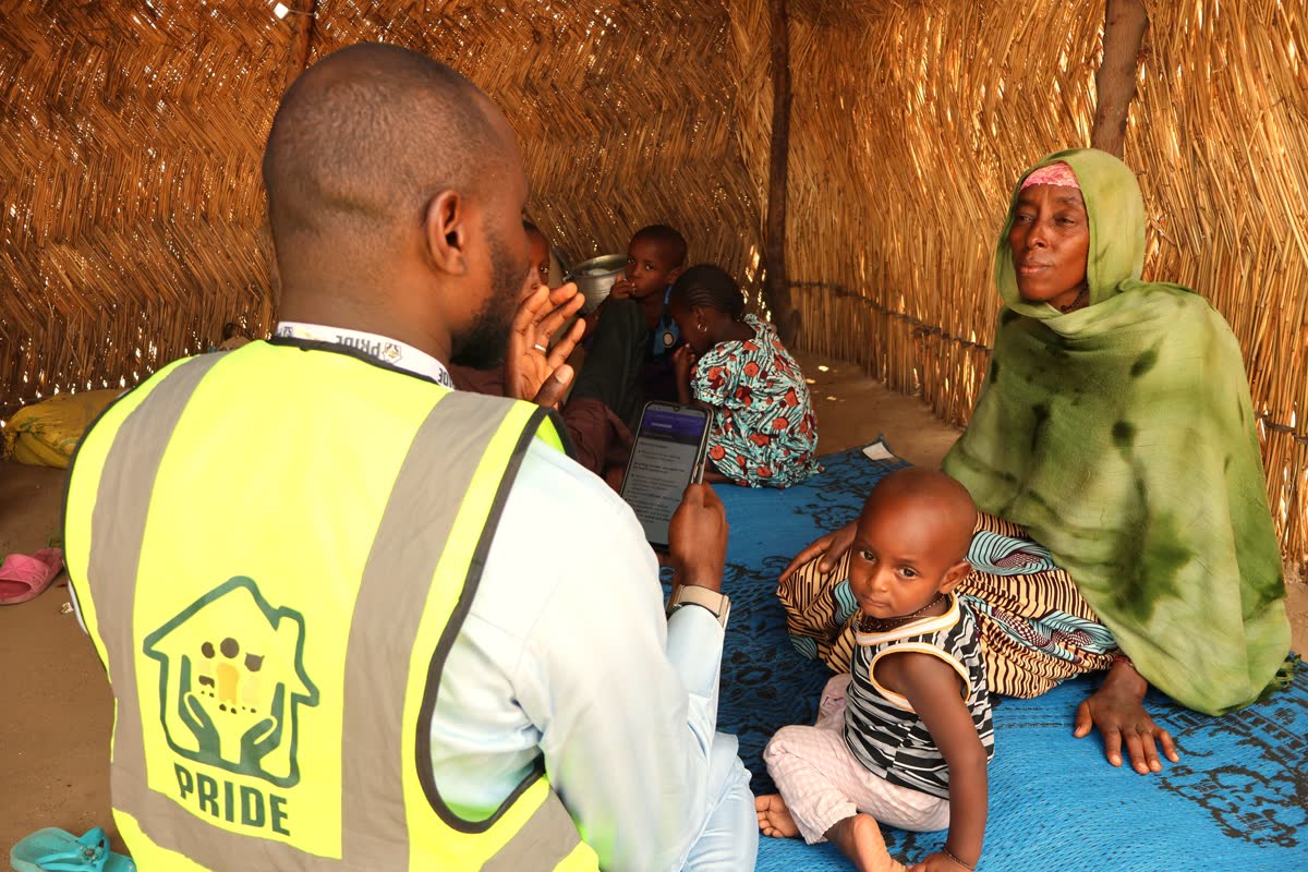 A PRIDE frontline worker in a hi-vis vest, seen from behind, holds a phone showing a Connect consent form while talking with a woman and four children sitting on a mat inside a thatched shelter.